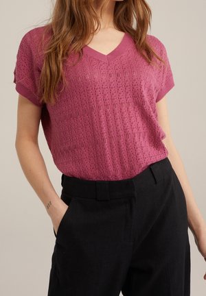 Růžový pletený top s výstřihem do V a krátkými rukávy, zdobený texturovaným vzorem. Doplněný černými kalhotami zvýrazňujícími detaily kapes.