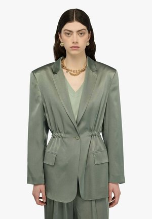Donna con lunghi capelli scuri che indossa un blazer di raso verde chiaro con vita aderente, pantaloni abbinati, collana a catena d'oro e orecchini a cerchio d'oro.