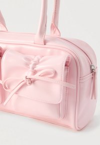 Bolso de mano de cuero sintético rosa con dos asas, un detalle de lazo en el frente y cierre de cremallera. Cuenta con dos bolsillos pequeños con solapas.