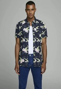 Homem a usar uma camisa de manga curta com padrão floral escuro sobre uma t-shirt branca e jeans azuis, encostado a um fundo cinza liso.