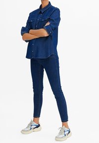Camisa de denim y jeans skinny en azul oscuro. La camisa tiene botones a presión y bolsillos en el pecho. Las zapatillas son blancas con detalles azules.