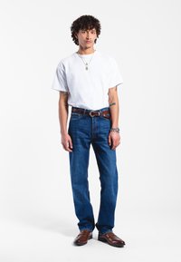 Camiseta blanca, jeans de mezclilla azul con corte recto, cinturón de piel marrón y mocasines marrones. La joyería incluye collares en capas y un reloj.