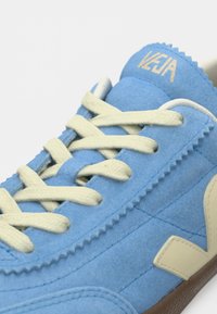 Απαλή μπλε σουέντ αθλητική τσόκα με κρεμ κορδόνια, μπεζ λεπτομέρεια λογότυπου και τη σήμανση "VEJA" στη γλώσσα, κοντινή άποψη.