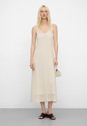 Blondynka w bezrękawnikowej beżowej dzianinowej maxi sukience, trzymająca małą kołową torebkę, stojąca na szarej podłodze na białym tle.