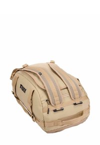 Sac de voyage beige avec des bretelles rembourrées, plusieurs poches zippées et un matériau synthétique durable. Il présente un logo sur le côté.