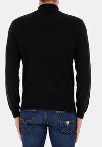 Pullover in maglia nera con collo alto, maniche lunghe e una texture liscia. Indossato con jeans blu caratterizzati da design distintivi delle tasche posteriori.