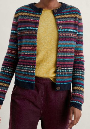 Personne portant un cardigan à boutons avec un motif multicolore sur un pull jaune et un pantalon en velours côtelé violet foncé, debout devant un fond uni.
