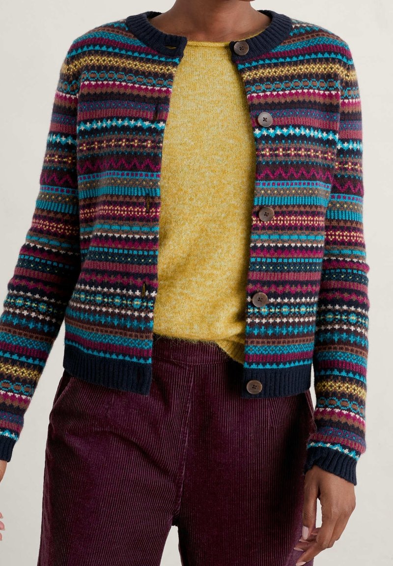 Personne portant un cardigan à boutons avec un motif multicolore sur un pull jaune et un pantalon en velours côtelé violet foncé, debout devant un fond uni.