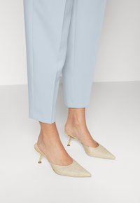 Beige pointed-toe mules made of textured material with gold accents and a stiletto heel, paired with light blue tapered trousers.