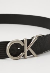 Fekete bőrszíj, dombornyomott mintázattal. Ezüstszínű fém csattal rendelkezik, amelyen a "CK" logó látható. Állítható, többlyukas méretmegoldással.