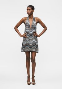 Γυναίκα στέκεται με τα χέρια στη μέση φορώντας αμάνικο μίνι φόρεμα με V-λαιμόκοψη και σχέδιο, καθώς και σανδάλια με ψηλό τακούνι σε απλό φόντο.