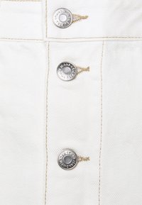 Jeans bianchi in denim con tre bottoni argentati e cuciture beige. Presentano una texture liscia e un design classico e dritto con cuciture visibili.