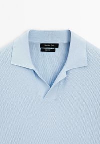Polo shirt in cotone azzurro chiaro con un motivo testurizzato, caratterizzato da un collo classico e un design a scollo a V. Etichetta del marchio visibile all'interno del collo.