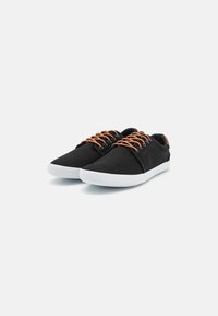 Zapatillas de lona negras con cordones marrones, suela de goma blanca, puntera redondeada y detalles de costura en contraste a los lados.