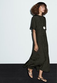Langes, fließendes olivgrünes Kleid aus strukturiertem Stoff, mit kurzen Ärmeln und entspannter Passform. Getragen mit schlichten schwarzen Sandalen und einer Anhänger-Halskette.