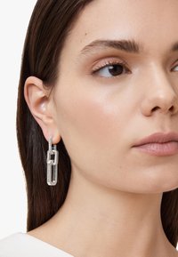 Orecchini in tonalità argento con un design geometrico, caratterizzati da link in acrilico trasparente e accenti di piccole pietre scintillanti. Texture liscia e forma moderna.