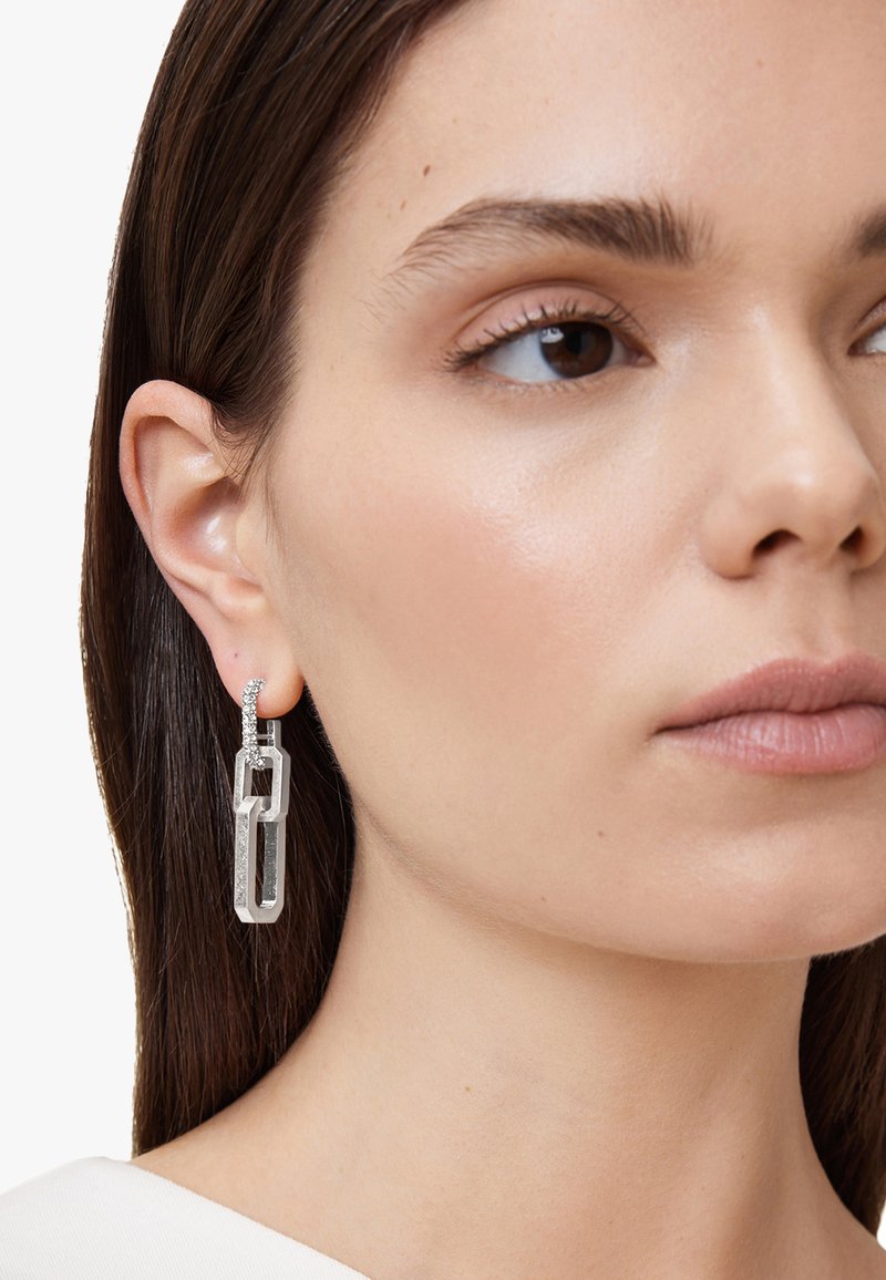 Orecchini in tonalità argento con un design geometrico, caratterizzati da link in acrilico trasparente e accenti di piccole pietre scintillanti. Texture liscia e forma moderna.