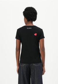Schwarzes Baumwoll-T-Shirt mit Rundhalsausschnitt, kurzen Ärmeln und einem roten Herzgrafik auf dem Rücken nahe dem Ausschnitt. Markenlogo über der Grafik.
