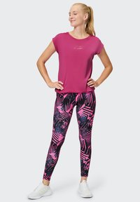 Magentafarbenes Kurzarm-Top mit „Venice Beach“-Aufdruck; gemusterte schwarze und pinke Leggings mit geometrischen Formen; weiße Sportschuhe.