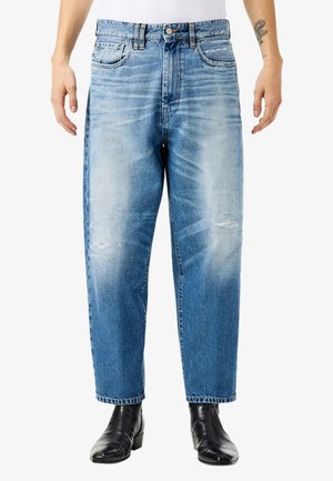 Uomo che indossa jeans azzurri sbiaditi con lievi segni di usura, stivali in pelle neri e una camicia bianca, in piedi con le braccia rilassate.