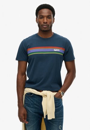Homme portant un t-shirt bleu marine avec des rayures rouges, violettes et vertes, un jean bleu, un pull beige noué autour de la taille et une montre-bracelet, debout devant un fond blanc.