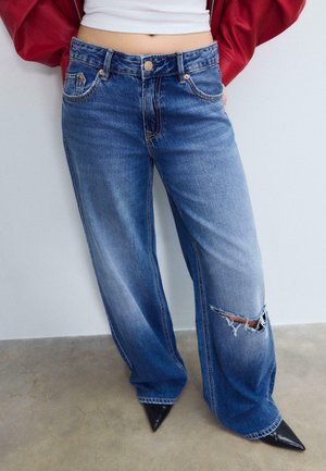 Osoba nosząca luźne niebieskie dżinsy z rozdarciem na prawym kolanie, czarne spiczaste buty, biały top i czerwoną kurtkę.