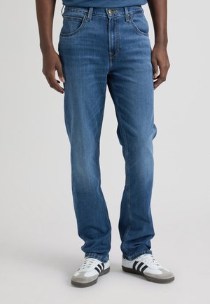 Person trägt mittelblaue gerade geschnittene Jeans und weiße Turnschuhe mit schwarzen Streifen, steht vor einem einfarbigen Hintergrund.