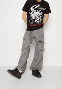 Camiseta de algodón negra con una impresión gráfica de una calavera y texto. Pantalones cargo grises con múltiples bolsillos. Zapatillas negras y marrones con suelas texturizadas.