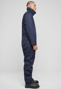 Tumši zils darba kombinezons ar augstu apkakli, ar rāvējslēdzēju un elastīgu jostu. Iezīmē garas piedurknes un rāvējslēdzējus pie apkakles. Redzamas melnas zābaki.