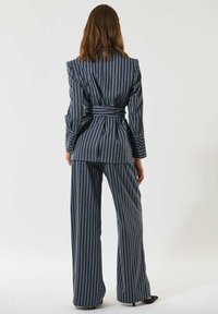 Räätälöity tummansininen viivoitettu puku vyötärönauhan kanssa, jossa on pitkät hihat ja leveälahkeiset housut. Sileä kangas, jolla on jämäkkä siluetti.