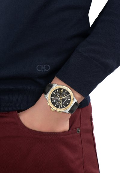 Čierno-zlaté hodinky s chronografom na ľavej ruke, oblečený v tmavomodrom svetri a bordových nohaviciach, ruka v vrecku.