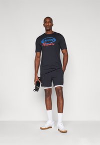 Sort t-shirt med blå og rød grafisk tekst, parret med sorte shorts med hvide kanter, hvide sokker og hvide sko. Holder en vandflaske.