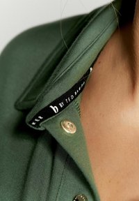 Camicia verde con colletto in tessuto testurizzato. Presenta un'etichetta nera con testo bianco e un bottone argentato al colletto. Dettaglio di chiusura visibile.