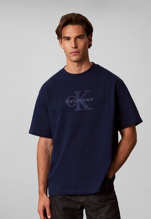 Ung mand iført en navyblå Calvin Klein t-shirt med korte ærmer og mørke jeans, stående mod en ensfarvet lys grå baggrund.