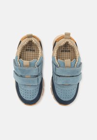 Blå mockasneakers med bruna accenter, som har två justerbara kardborreband, rund tå och en strukturerad gummisula. Inuti finns ett rutigt mönster.