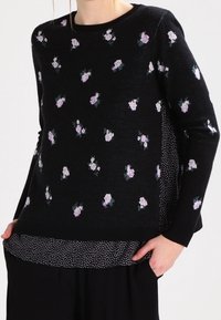 Pull noir avec un motif floral en nuances de rose et de vert, doté d'un ourlet à pois et de manches longues. Texture douce, col rond.