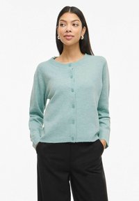 Gaiši tirkīza krāsas kardigans ar apaļu kakla izgriezumu, pogu aizdari priekšpusē, garām piedurknēm un maigu tekstūru. Stils melnās biksēs kontrastam.