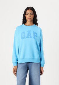 Sudadera de cuello redondo en azul claro, fabricada con tejido suave, que presenta un logo elevado de "GAP" en un azul ligeramente más oscuro. Corte holgado y puños acanalados.