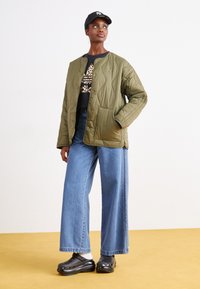 Olivno zelena prešita jakna s gumbi na pritisk, v kombinaciji z črnim grafičnim puloverjem in širokimi modrimi kavbojkami. Črni klobuki dopolnjujejo obleko.