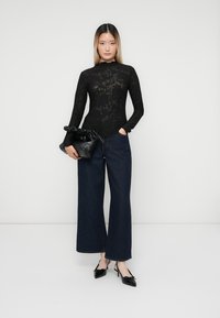 Černý krajkový bodysuit s dlouhými rukávy a květinovým vzorem, doplněný o tmavě modré široké kalhoty s vysokým pasem. Drží černou texturovanou kabelku.
