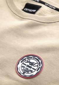 Beige sweatshirt met een gestructureerde afwerking, voorzien van een ronde patch met zwarte, witte en rode details, met de tekst "Sport and Co. 1985."
