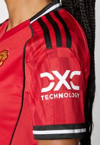 Röd sporttröja med svarta horisontella ränder på axlarna, ett strukturerat tyg och en framträdande vit logotyp med texten "DXC TECHNOLOGY" på ärmen.
