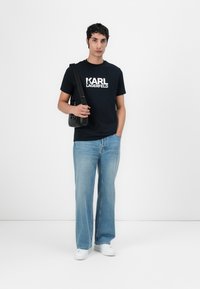 Marineblå kortermet T-skjorte med hvit "KARL LAGERFELD" tekst, kombinert med lyseblå vide jeans og hvite joggesko. Svart crossover-veske inkludert.