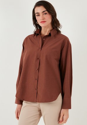 Bruine blouse met lange mouwen, voorzien van een zachte textuur en een afgeronde zoom. Gekleed met beige broeken.