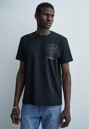 Mężczyzna ubrany w czarną koszulkę Off-White z białym logo na piersi, niebieskie dżinsy, stojący na tle jednolitego jasnoszarego tła.