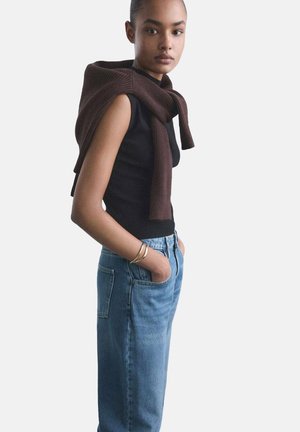 Black sleeveless top paired with blue high-waisted jeans. A brown knit sweater is draped over the shoulders. Simple silver bracelets on wrist.