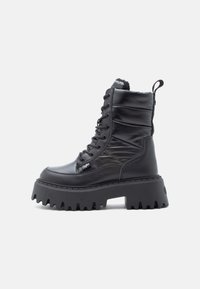 Μαύρο μποτάκι με ψηλό τον αστράγαλο, με κορδόνια, που διαθέτει παχύ αντιολισθητικό πάτο, επάνω μέρος με επένδυση και ταπακέ διεπιφάνειας, και ετικέτα μάρκας στο πλάι.