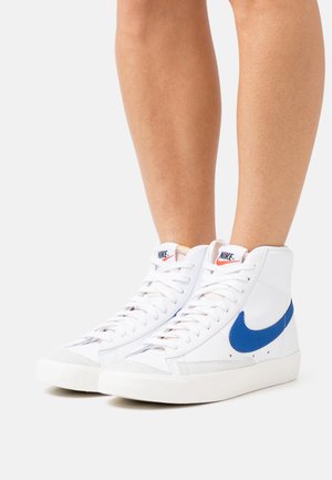 Höga sneakers i vitt läder med blå Nike-swoosh, gummisula, rund tå och tydligt skosnörningssystem.