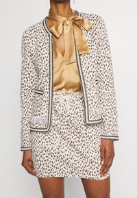 Leopardi-kuosiset bleiseri ja mini-hame setti valkoisena ja ruskeana mustilla yksityiskohdilla. Sisältää silkkisen nauhakiinnitteisen paidan ja fringed yksityiskohdat taskuissa.