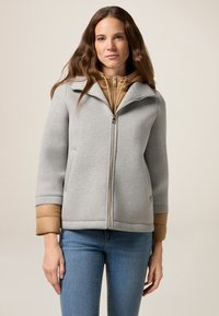 Giacca grigia con zip, colletto e dettagli beige sui polsini, caratterizzata da una texture liscia e da un design strutturato. Indossata sopra uno strato con cappuccio marrone.